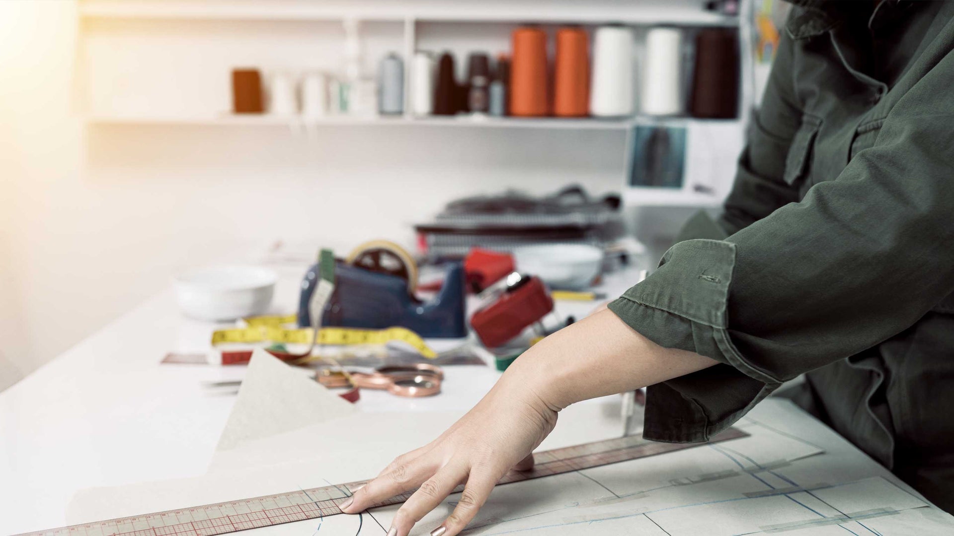A person's hand and arm are shown using a metal ruler on a sheet of pattern paper on a white table, with sewing and crafting tools like scissors, tape, and spools of thread in a blurred workshop background.