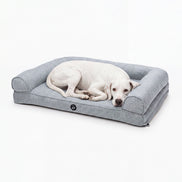 A white dog lies resting on a light gray fabric pet bed with a raised bolster border, photographed against a clean white studio background.